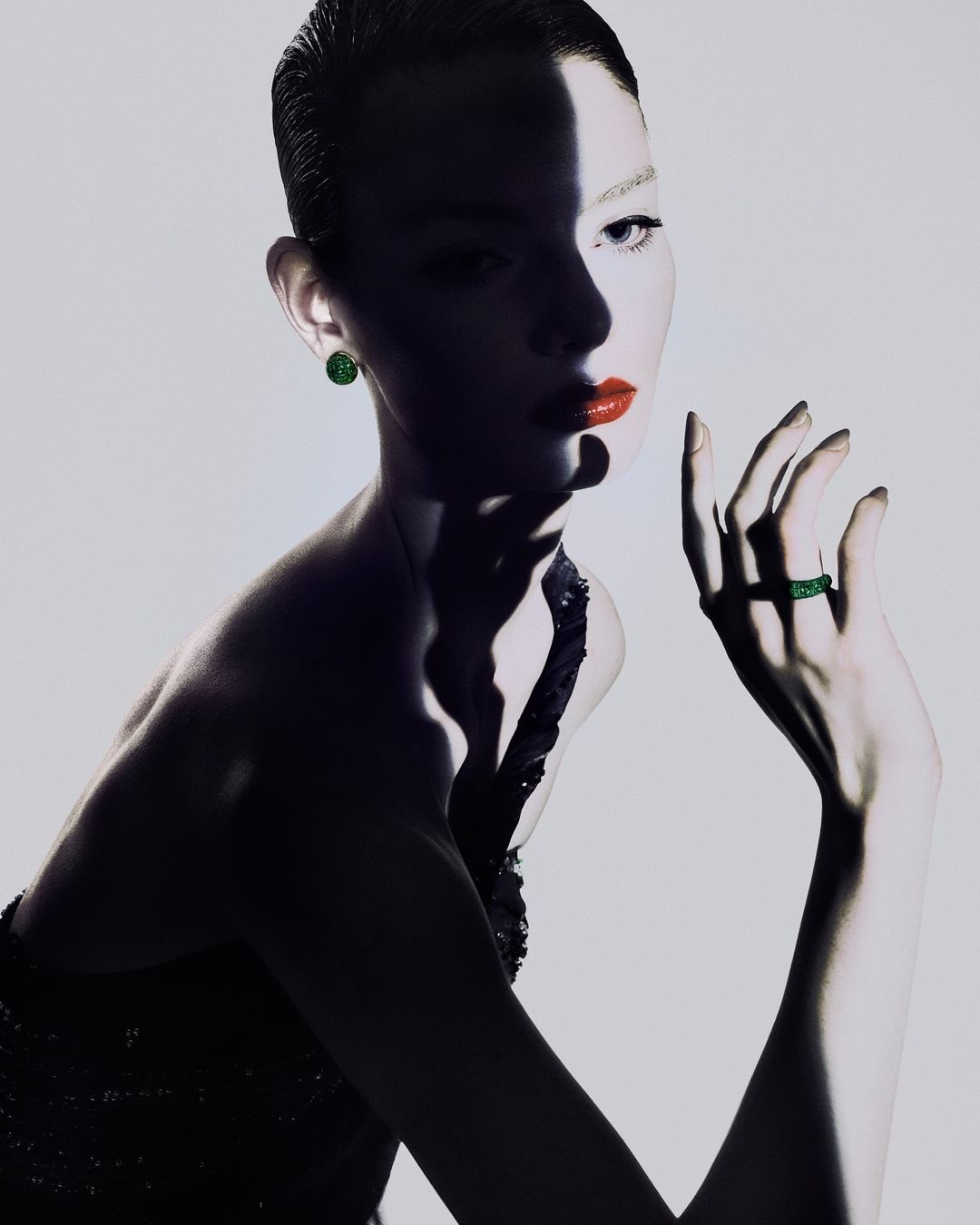 Close-up studio shot of a fashionable adult woman in an elegant black dress and green jewelry, with dramatic lighting and makeup.