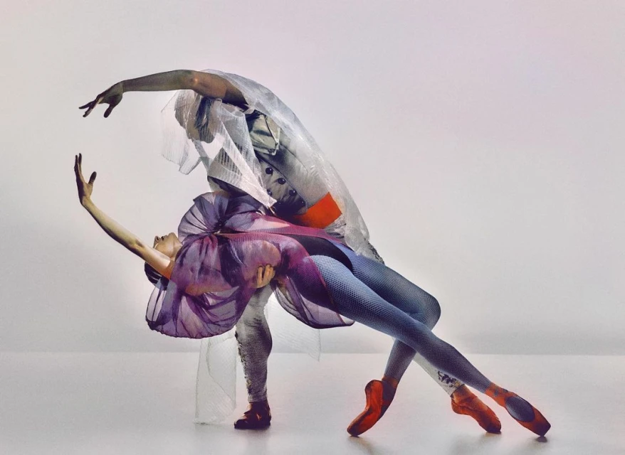 Two ballet dancers in a studio. One dancer holds the other in a pose with artistic costumes and pointe shoes, on a white background.