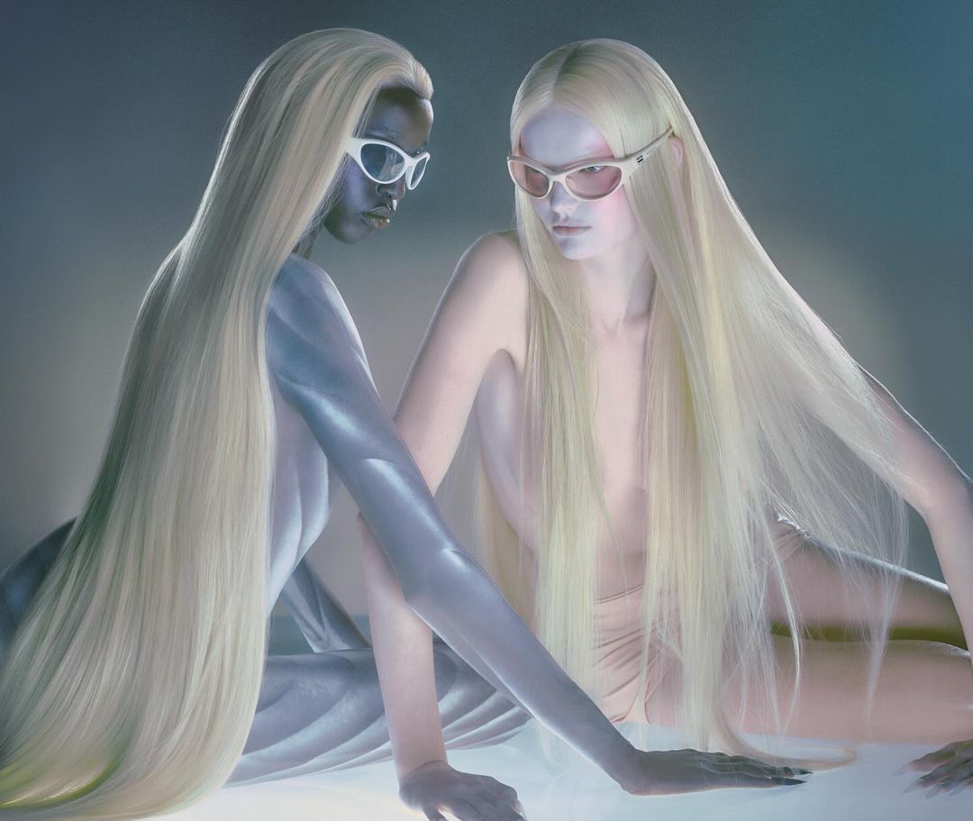Two fashion models with long blonde hair pose in sunglasses against a blue background, creating an ethereal and modern aesthetic.