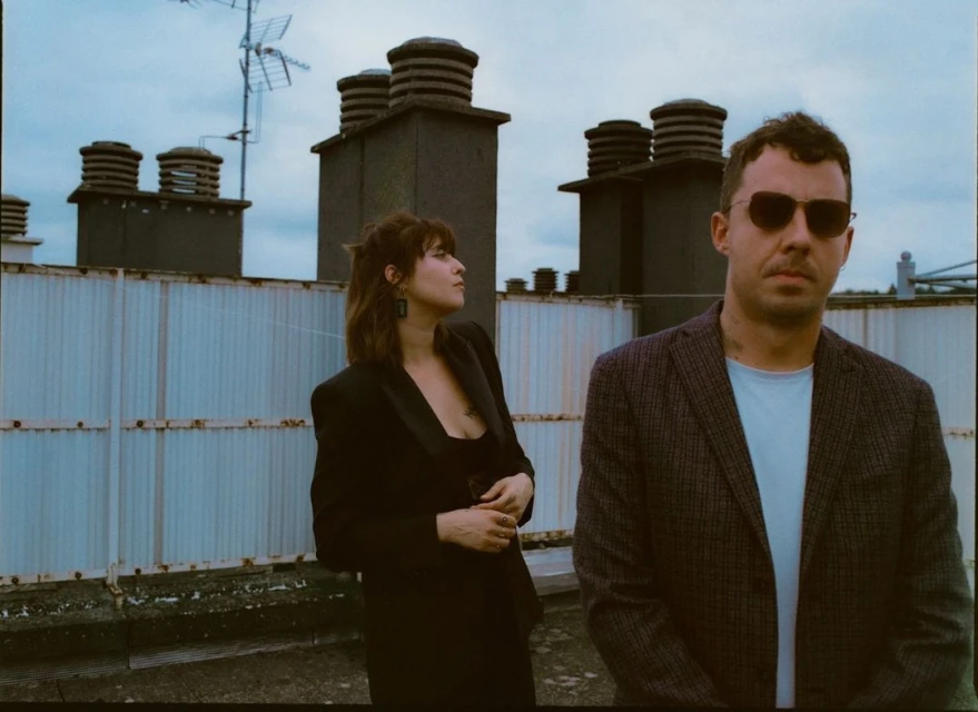 Two adults pose on a rooftop with city chimneys and cloudy sky. The woman wears a black blazer and the man wears a grey plaid blazer.