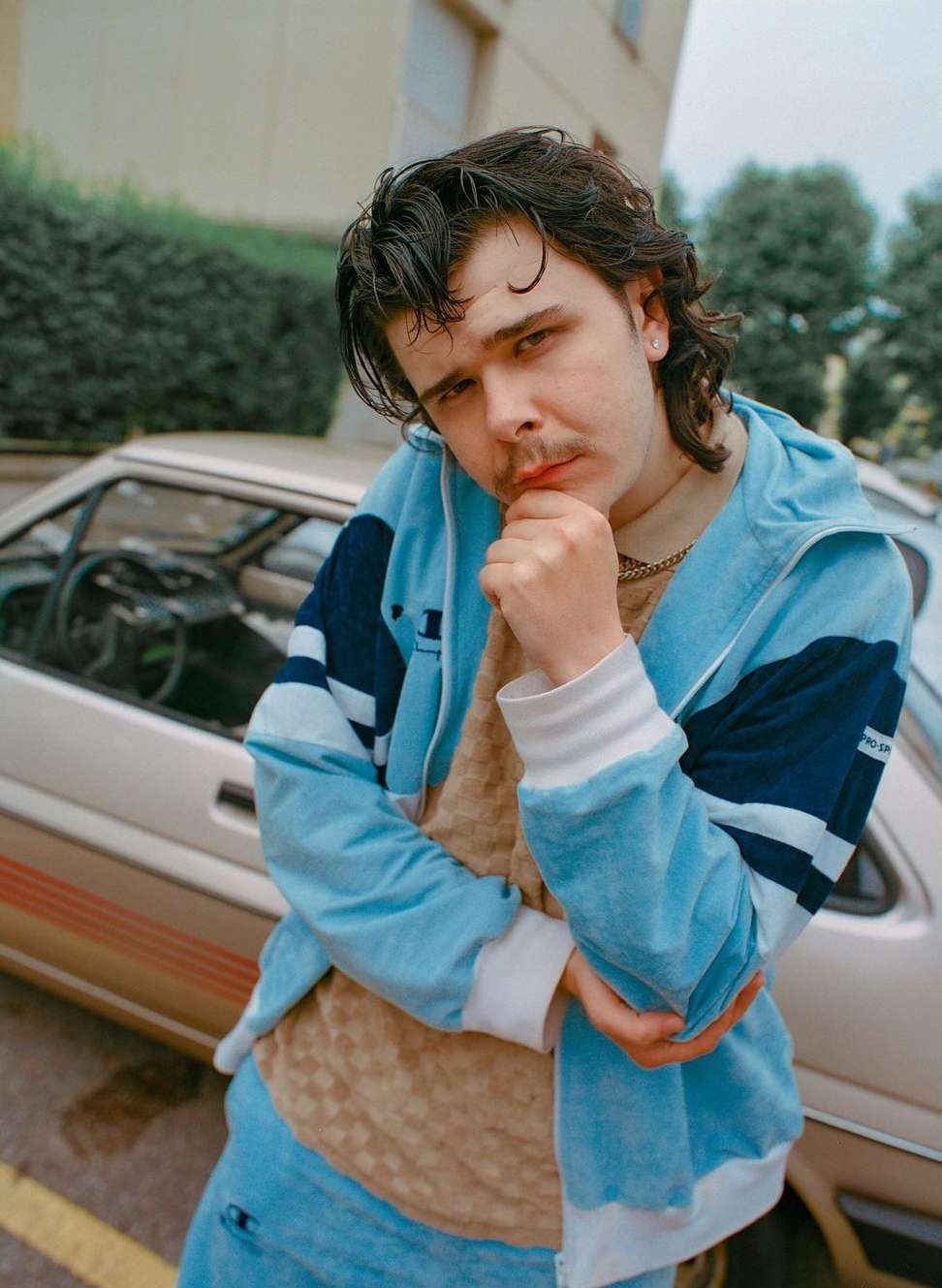 Portrait of a young adult male wearing a light blue tracksuit jacket and jeans, posing near a tan car in an outdoor urban setting.