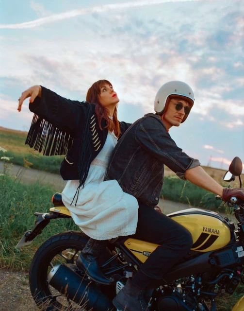 Couple riding a yellow Yamaha motorcycle on a rural road under a bright sky. Woman in white dress, man in helmet and denim.