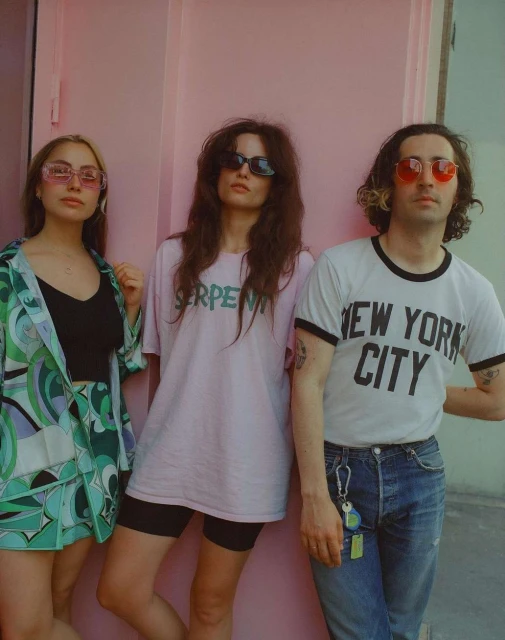 Three people stand in front of a pink wall wearing t-shirts and sunglasses. The women wear fashionable attire, and the man wears jeans.