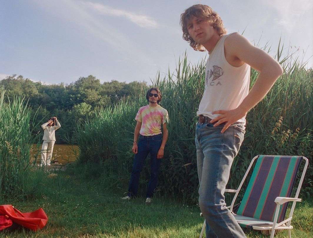 Three young men are standing in a grassy field next to a pond, one in a white tank top, blue jeans and another in tie dye shirt.