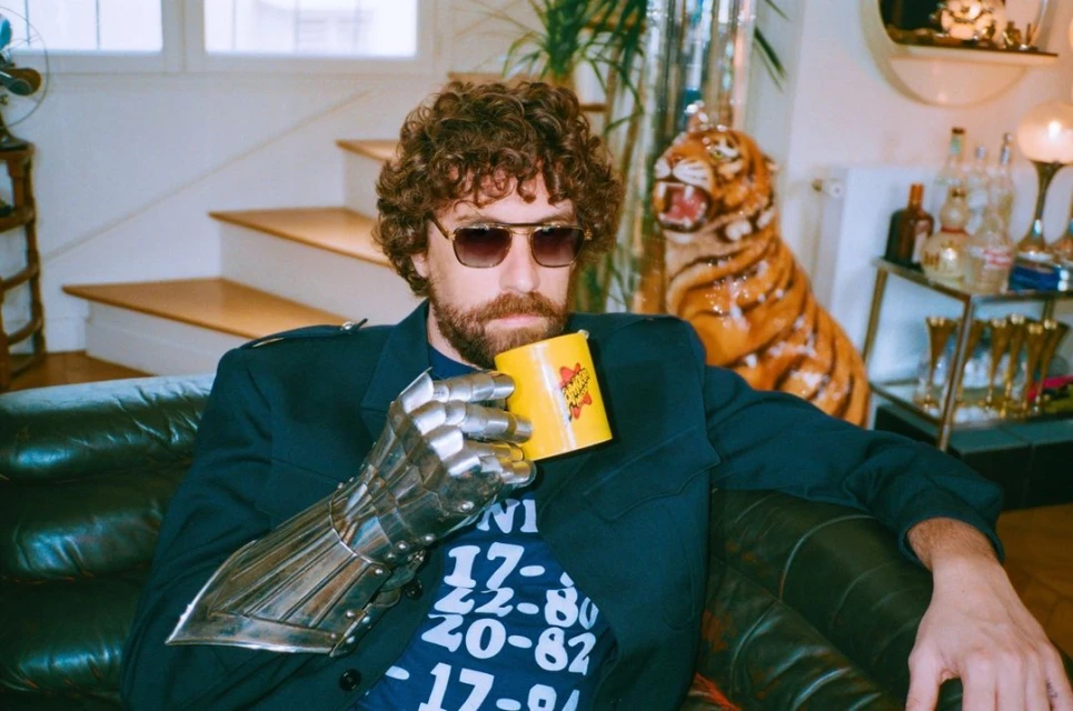 Adult male with curly hair and sunglasses reclines indoors, holding a yellow mug, wearing a blazer and a silver metal arm covering.