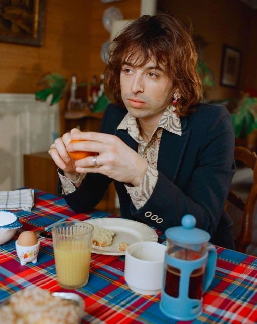 Man sits at a breakfast table with a patterned tablecloth, holding an orange. There is a French press, toast, and juice on the table.