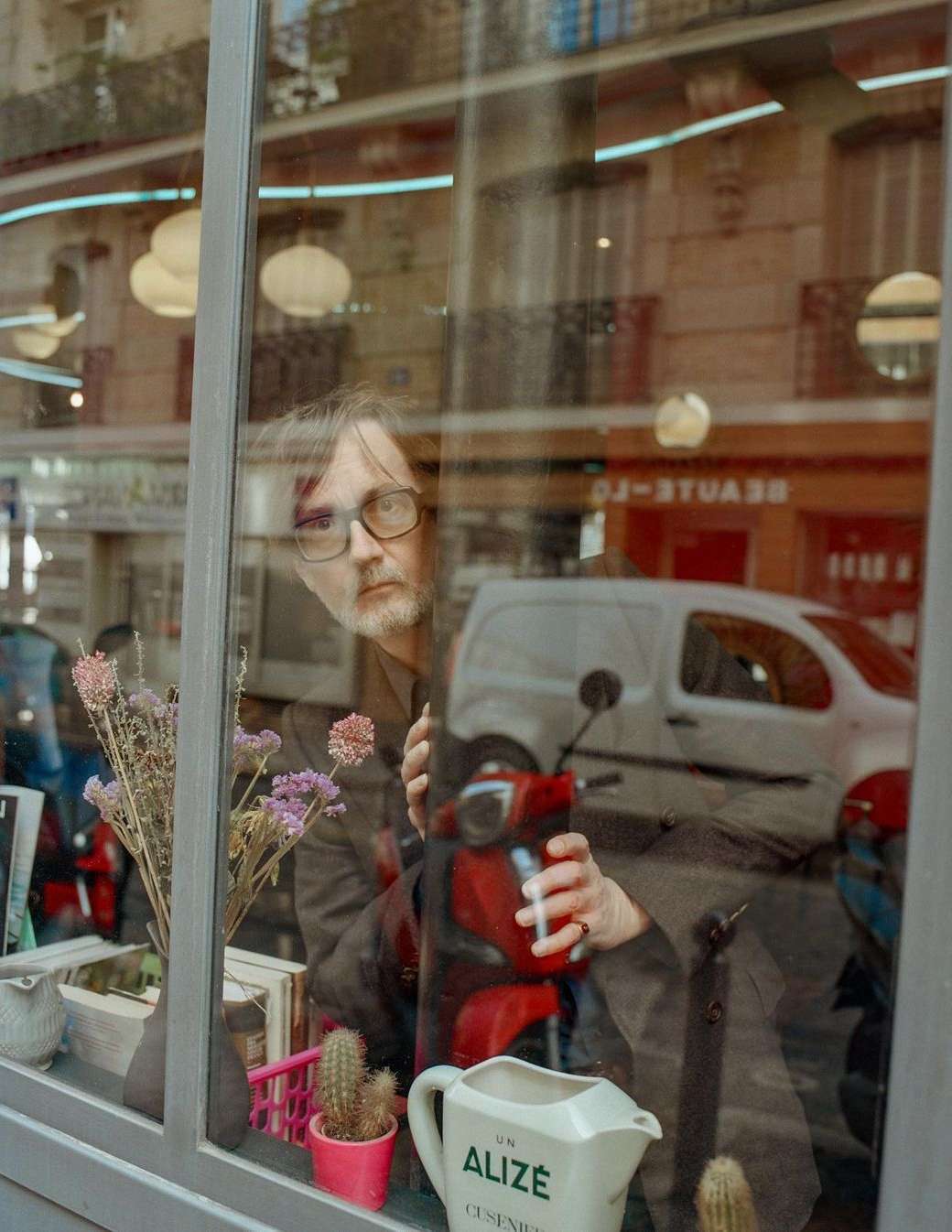 Man looking through a window in a gray jacket, reflected in glass with a red scooter, plants, and a street scene in Paris.
