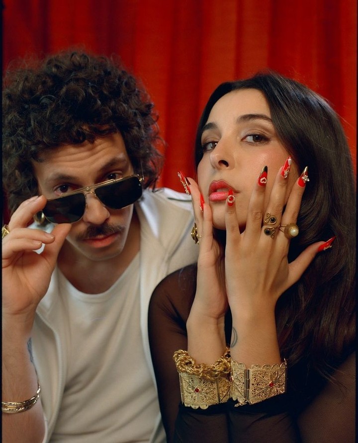 A stylish couple poses indoors against a red curtain backdrop, showcasing bold fashion choices like gold jewelry and sunglasses.