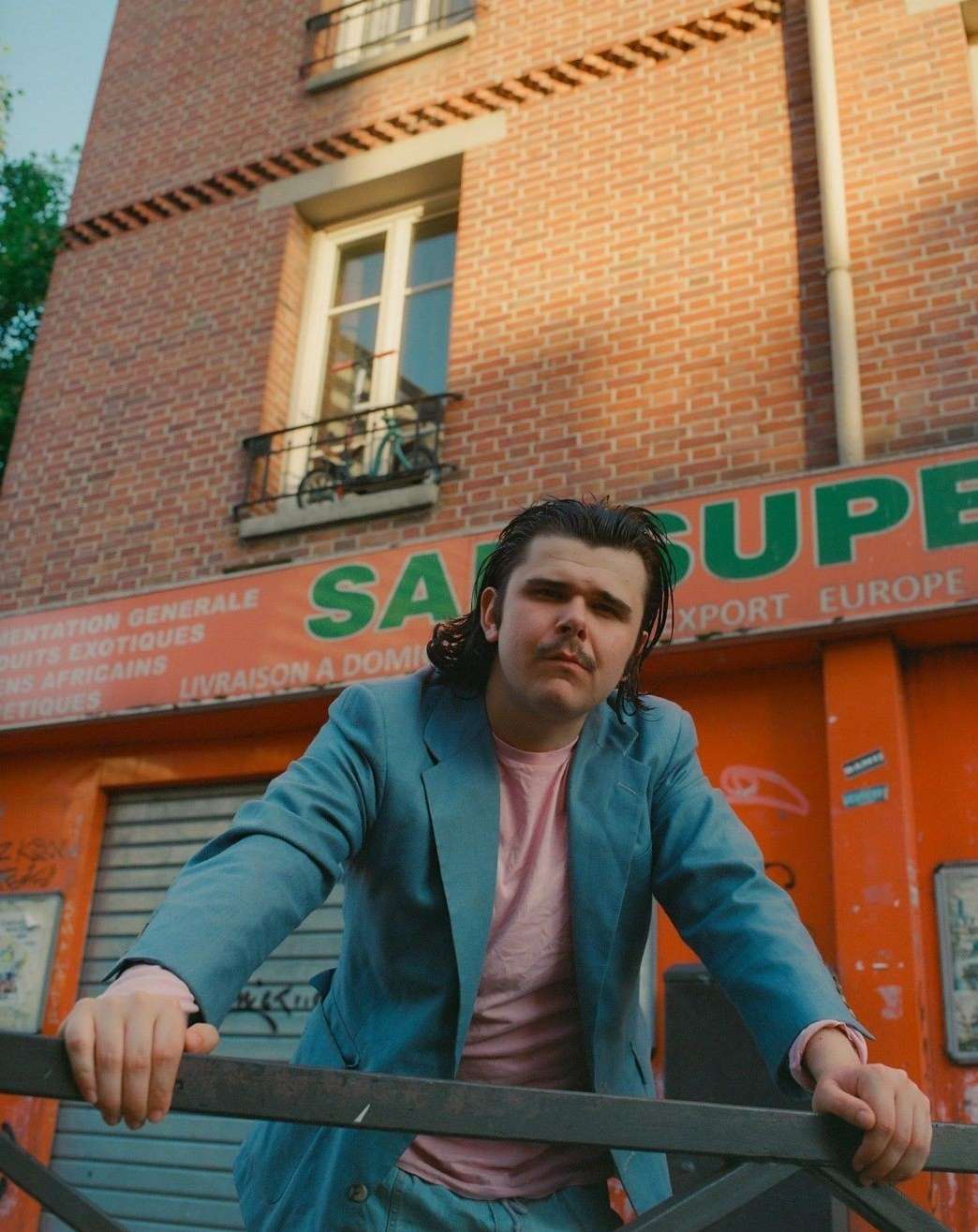 Young man in blue suit poses outdoors in front of brick building with shop. Portrait shot, medium angle, natural light, pink shirt.