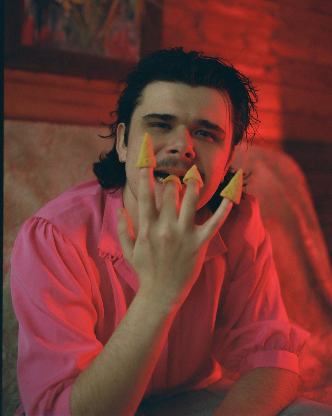 Man poses with snack chips on fingers, wearing a pink shirt, with red lighting in an indoor environment.