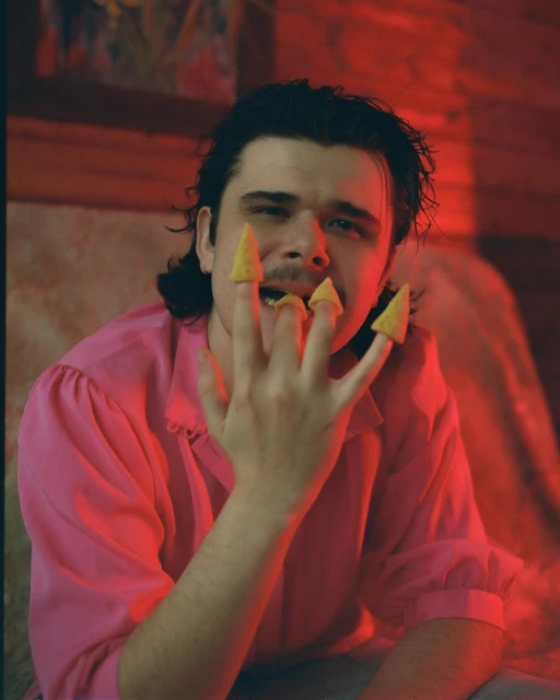 Man poses with snack chips on fingers, wearing a pink shirt, with red lighting in an indoor environment.