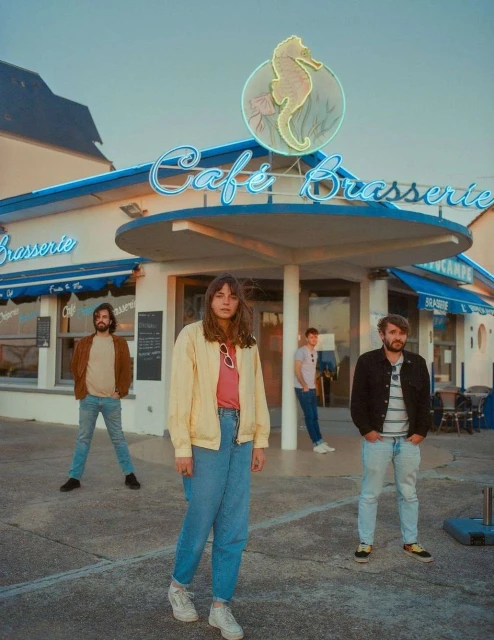Four people stand outside a cafe with a seahorse neon sign wearing casual clothing in a medium shot on a sunny day.