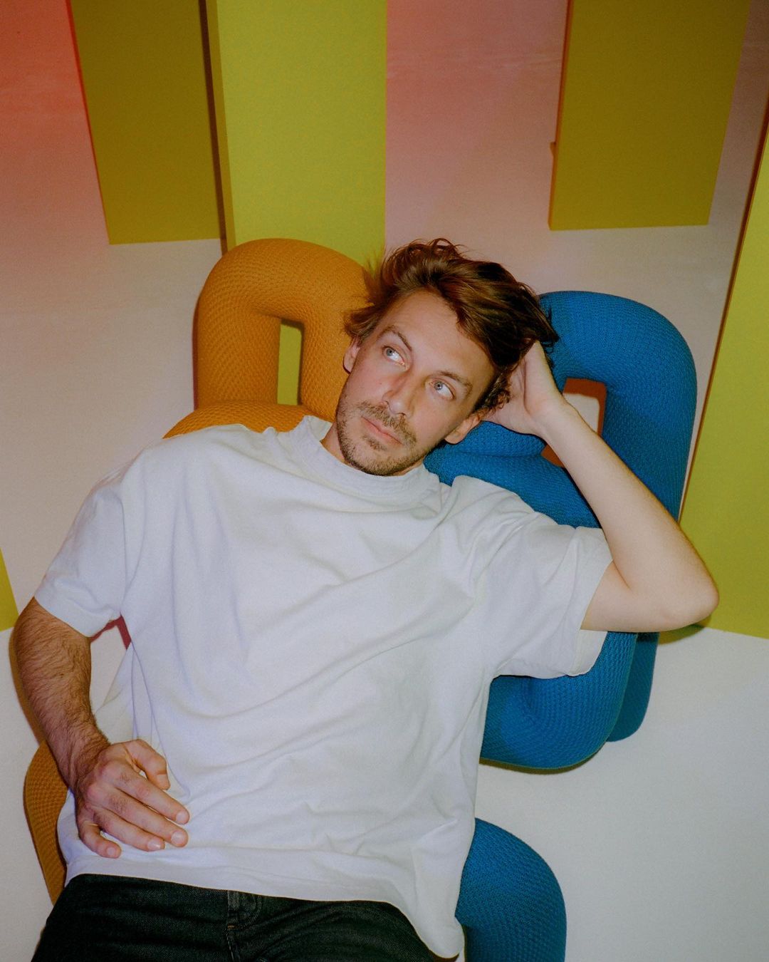 Indoor portrait of a Caucasian man in a white t-shirt, resting against a colorful wall structure, looking upwards with a relaxed expression.