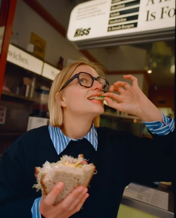 Blond woman in glasses eating a sandwich in a cafe setting, showcasing a blend of casual fashion and everyday food indulgence.