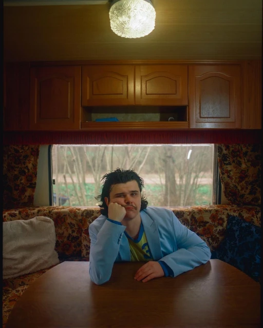 Adult male sitting inside campervan with brown cabinets, table, floral sofa, and trees visible through the window in background.