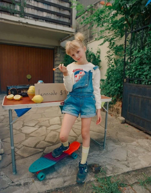 Young girl stands at a lemonade stand outside her home, next to a skateboard, selling lemonade for one dollar on a sunny day.