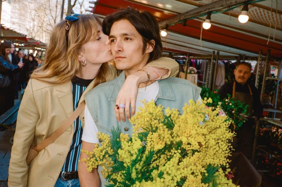 Woman kisses man holding yellow flowers at a market. The couple wears casual clothing in an outdoor urban setting.