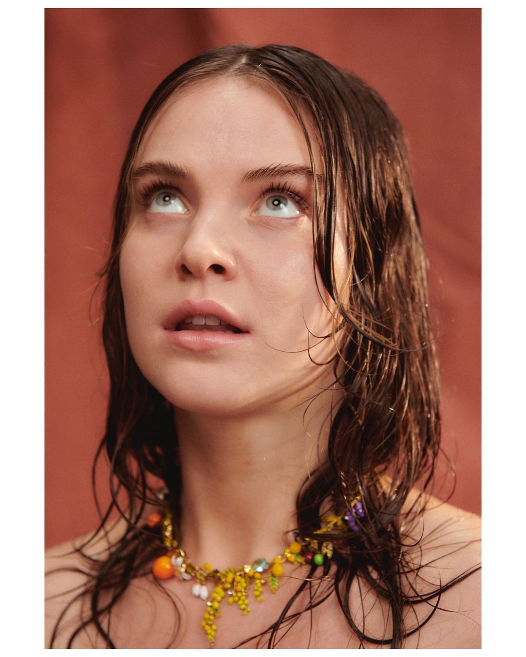 Portrait of a young adult female with wet brown hair, looking up with a colorful beaded necklace against an orange background.
