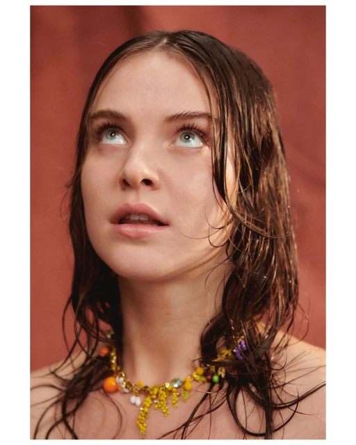 Portrait of a young adult female with wet brown hair, looking up with a colorful beaded necklace against an orange background.