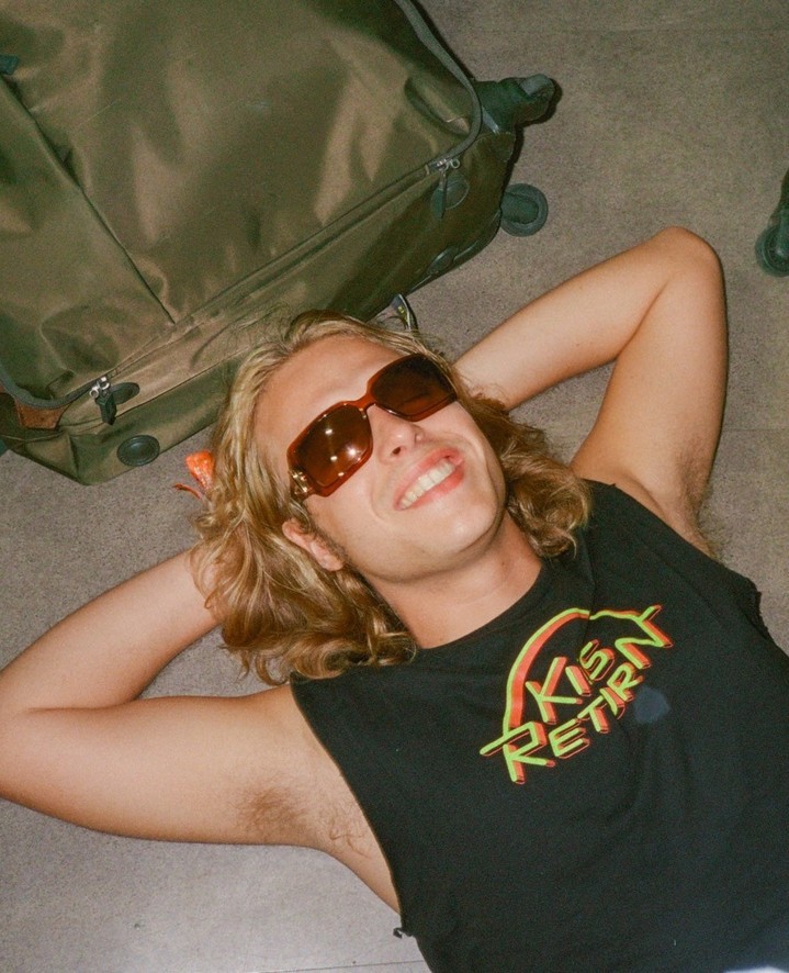 Young adult male with sunglasses lying down on a floor with a suitcase, smiling at the camera with a t-shirt in an indoor setting.
