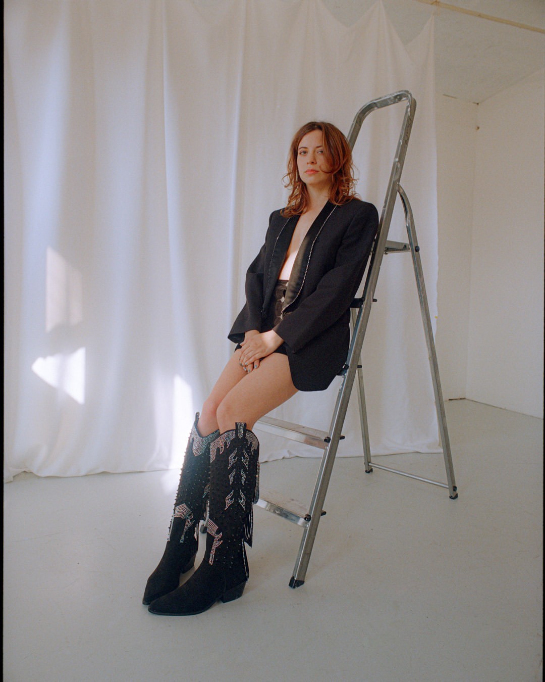 A woman in a blazer and cowboy boots is sitting on a stepladder in a photo studio, the image has natural light and white background.