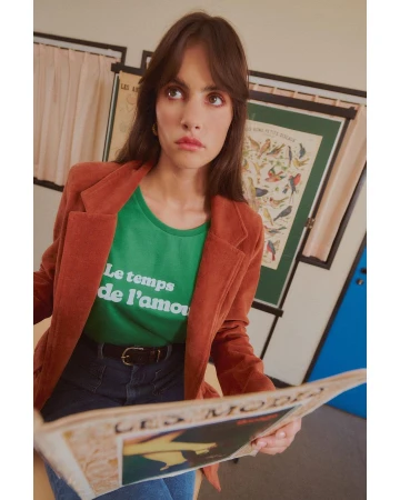 Adult female in a rust-colored blazer over a green t-shirt, reading a newspaper indoors near a window with natural light.