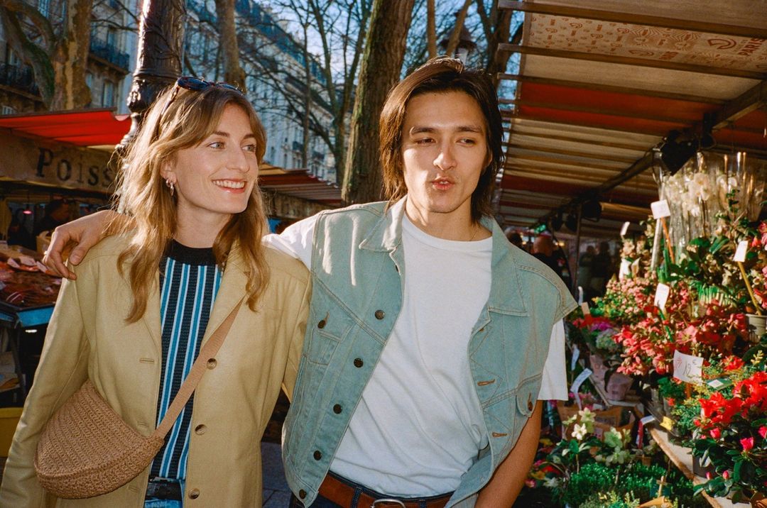 Woman and man stroll through a flower market in Paris, France, wearing casual jackets and smiling in natural daylight.
