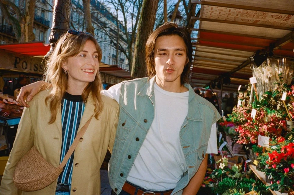 Woman and man stroll through a flower market in Paris, France, wearing casual jackets and smiling in natural daylight.