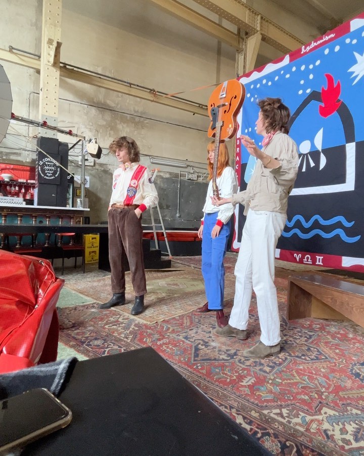 Three people standing in front of a painted backdrop in a loft-style space with a red chair, patterned rug, and natural light.