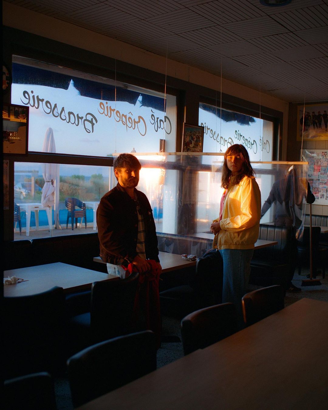 Two people stand inside a cafe with large windows overlooking an outdoor seating area and beach at sunset with orange lighting.