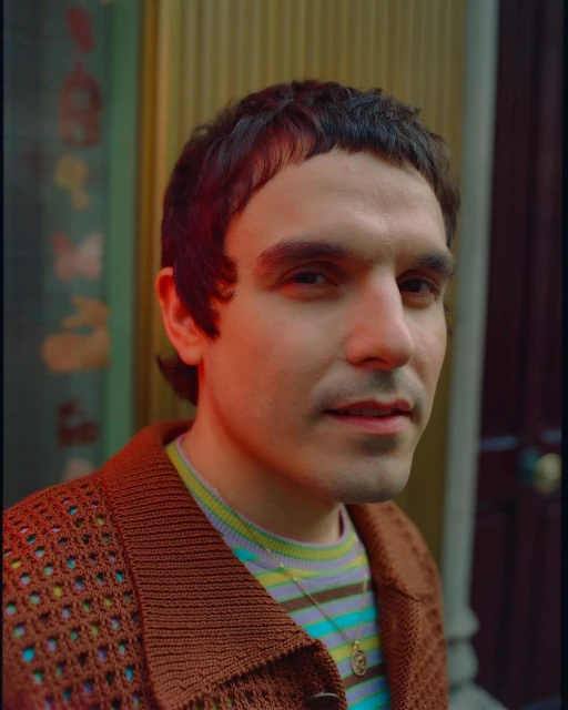 Portrait of a young adult male with short dark hair in a striped shirt and brown cardigan, standing indoors against a gold background.