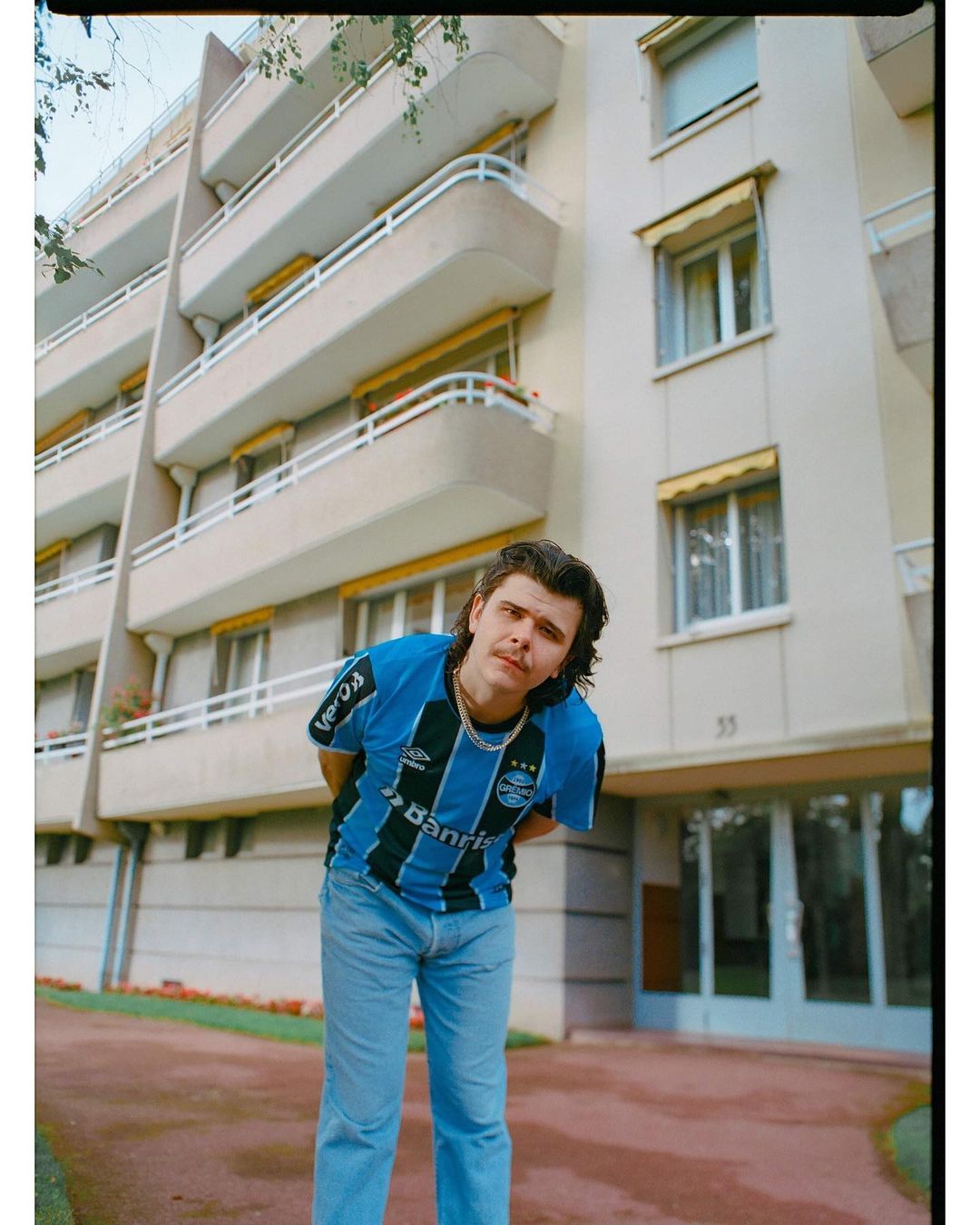 Young adult man poses in a soccer jersey and jeans against a beige apartment building with balconies on a sunny day outdoors.