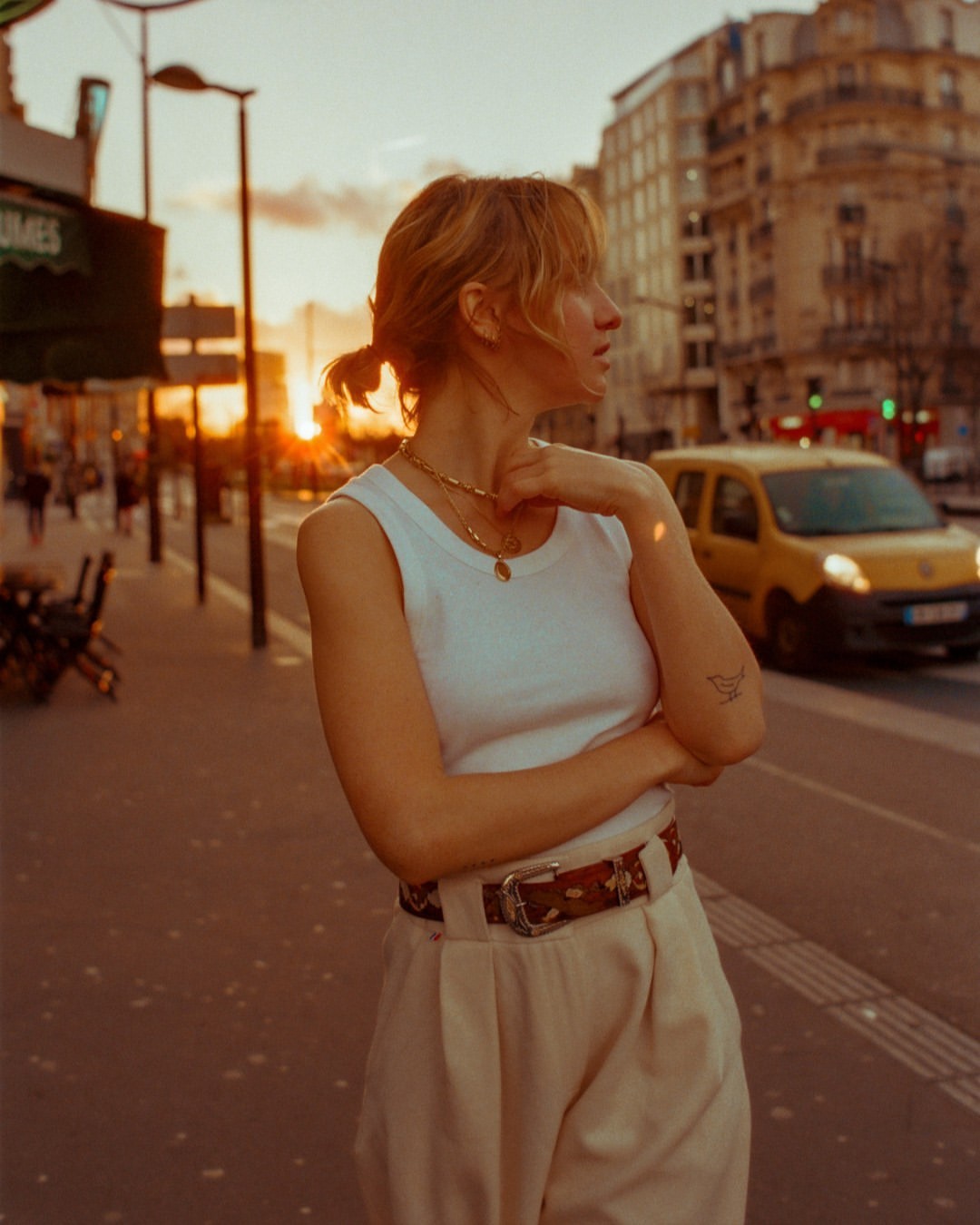 A woman in a white tank top and trousers stands on a city street at sunset, with buildings and a yellow van in the background.