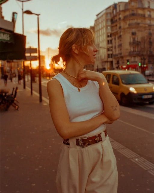 A woman in a white tank top and trousers stands on a city street at sunset, with buildings and a yellow van in the background.