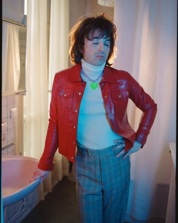 Portrait of a young adult with dark hair wearing a red leather jacket, a white turtleneck, and plaid pants in a bathroom.