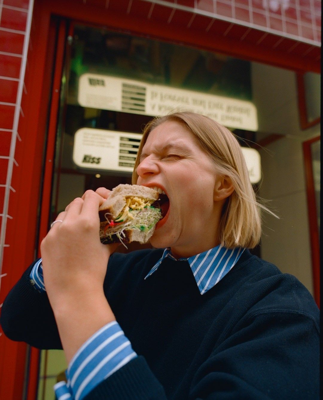 Woman takes a bite of a large sandwich filled with salad in front of a red-tiled building with a menu board in the background.