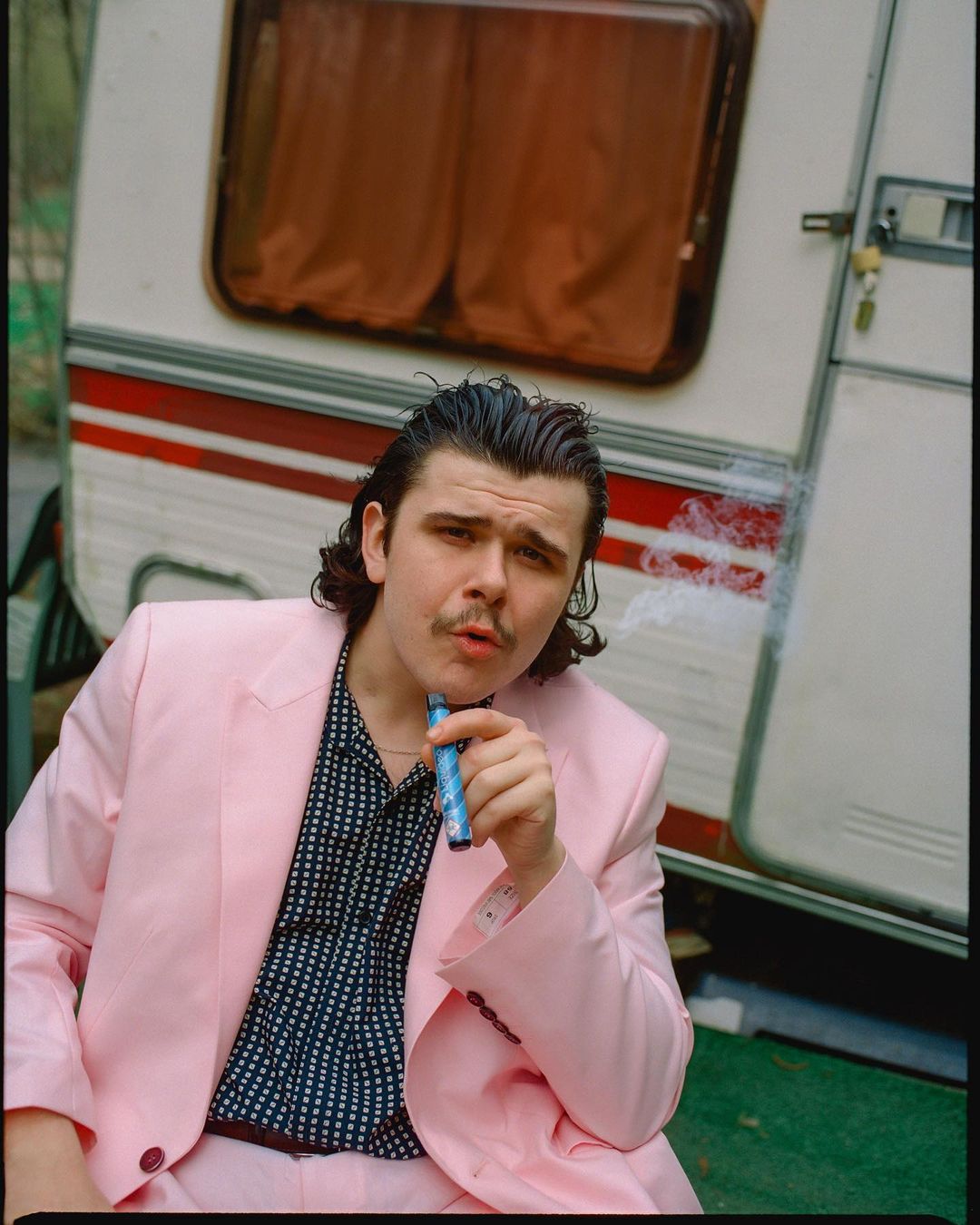 Portrait of a young adult male in a pink suit smoking an e-cigarette in front of a vintage camper trailer outdoors during daytime.