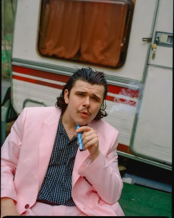 Portrait of a young adult male in a pink suit smoking an e-cigarette in front of a vintage camper trailer outdoors during daytime.