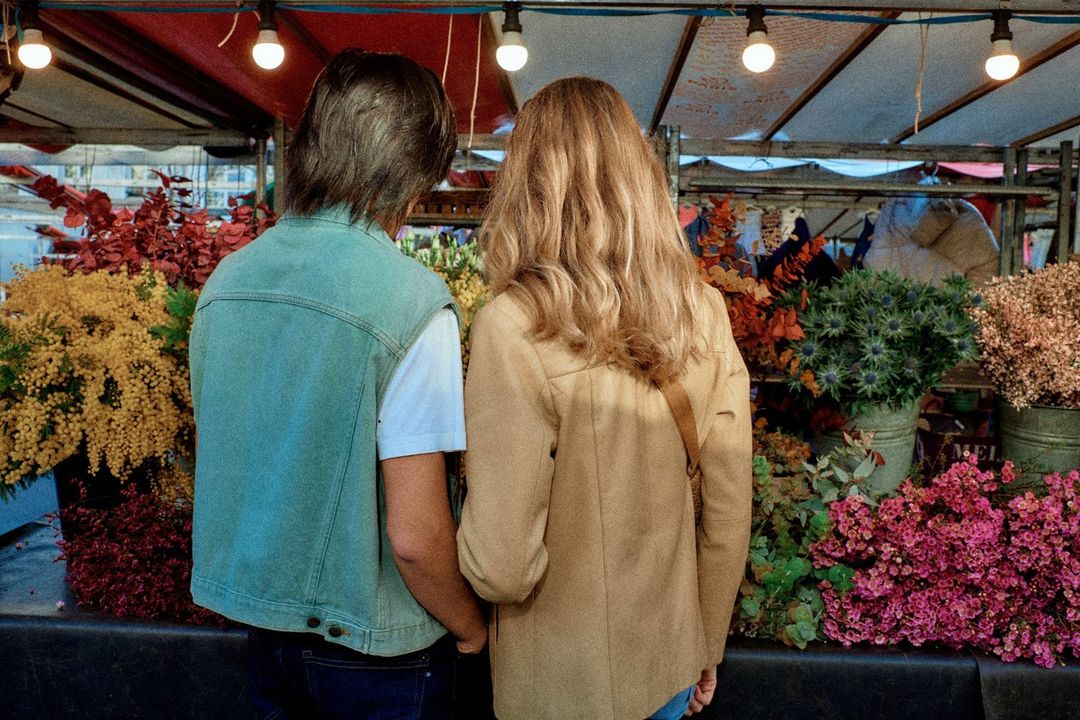 Back view of a couple standing at an outdoor flower market, featuring vibrant flowers, overhead lighting, and casual attire in daylight.