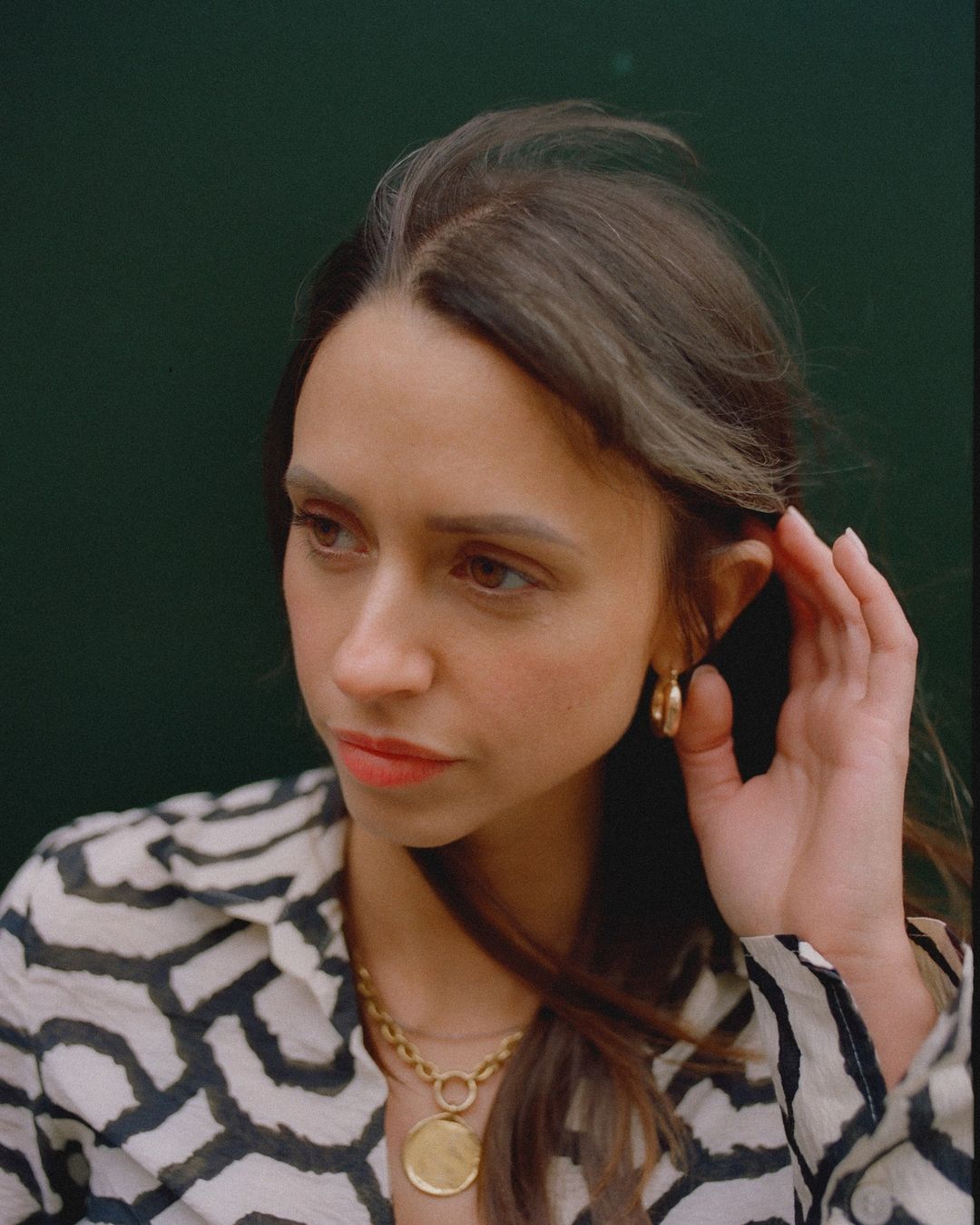 Portrait of a brunette woman with a patterned top and gold jewelry against a dark green backdrop, looking away with a thoughtful expression.