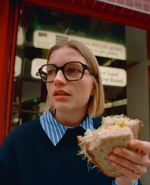 Woman in eyeglasses eating a large sandwich with sprouts and other ingredients in front of a shop with red trim.