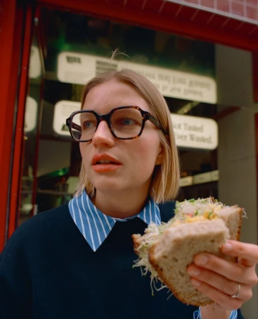 Woman in eyeglasses eating a large sandwich with sprouts and other ingredients in front of a shop with red trim.