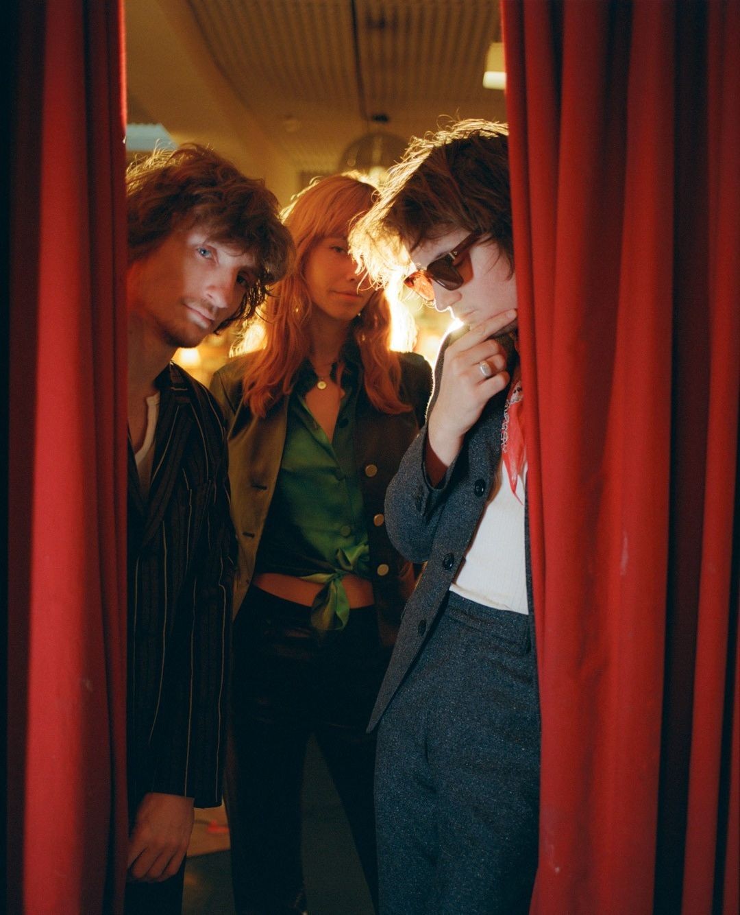 Three fashionably dressed individuals posing between red curtains in a warm, indoor setting with retro styling and soft, diffused lighting.