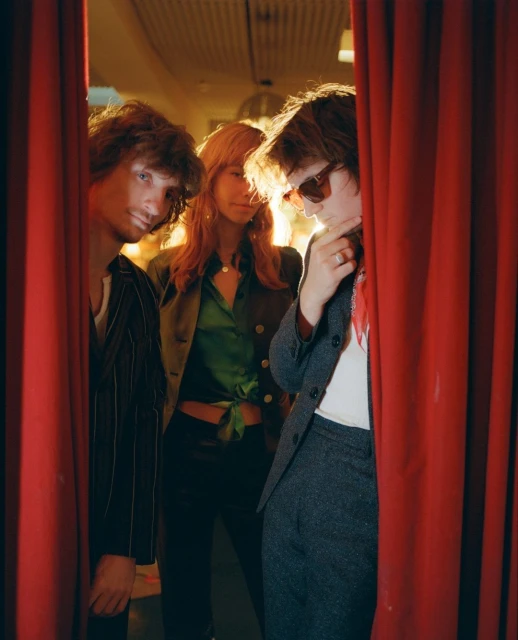 Three fashionably dressed individuals posing between red curtains in a warm, indoor setting with retro styling and soft, diffused lighting.