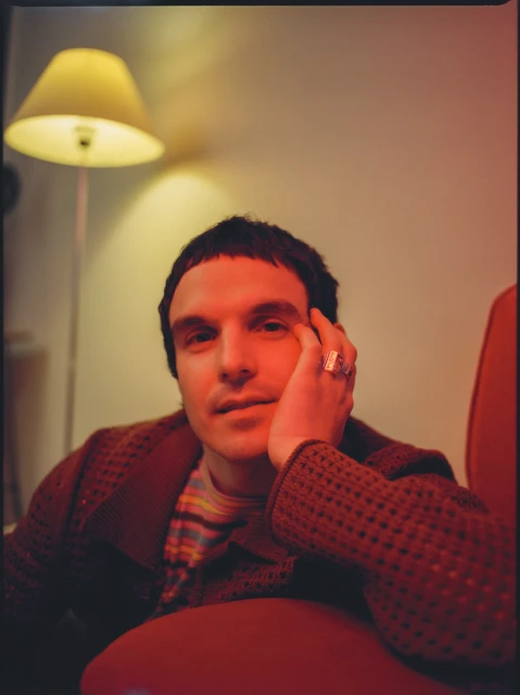 Portrait of a young adult man in a red chair with a yellow lamp in the background wearing a brown cardigan.