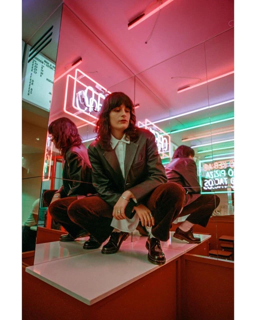 Stylish young woman in dark blazer crouching on a mirrored shelf in front of neon signs in an indoor setting.