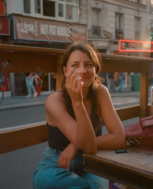 A woman seated at an outdoor table in an urban setting, smiling and holding a cigarette, with shops visible in the background.