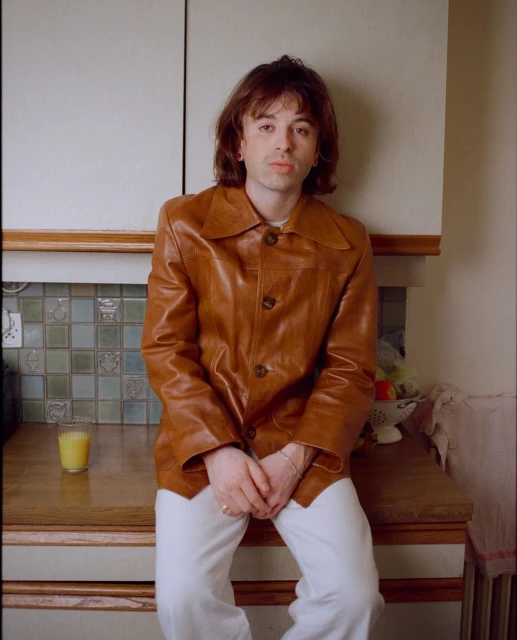 Adult male in a brown leather jacket posing in a kitchen with a glass of juice. Portrait captures indoor fashion and lifestyle.