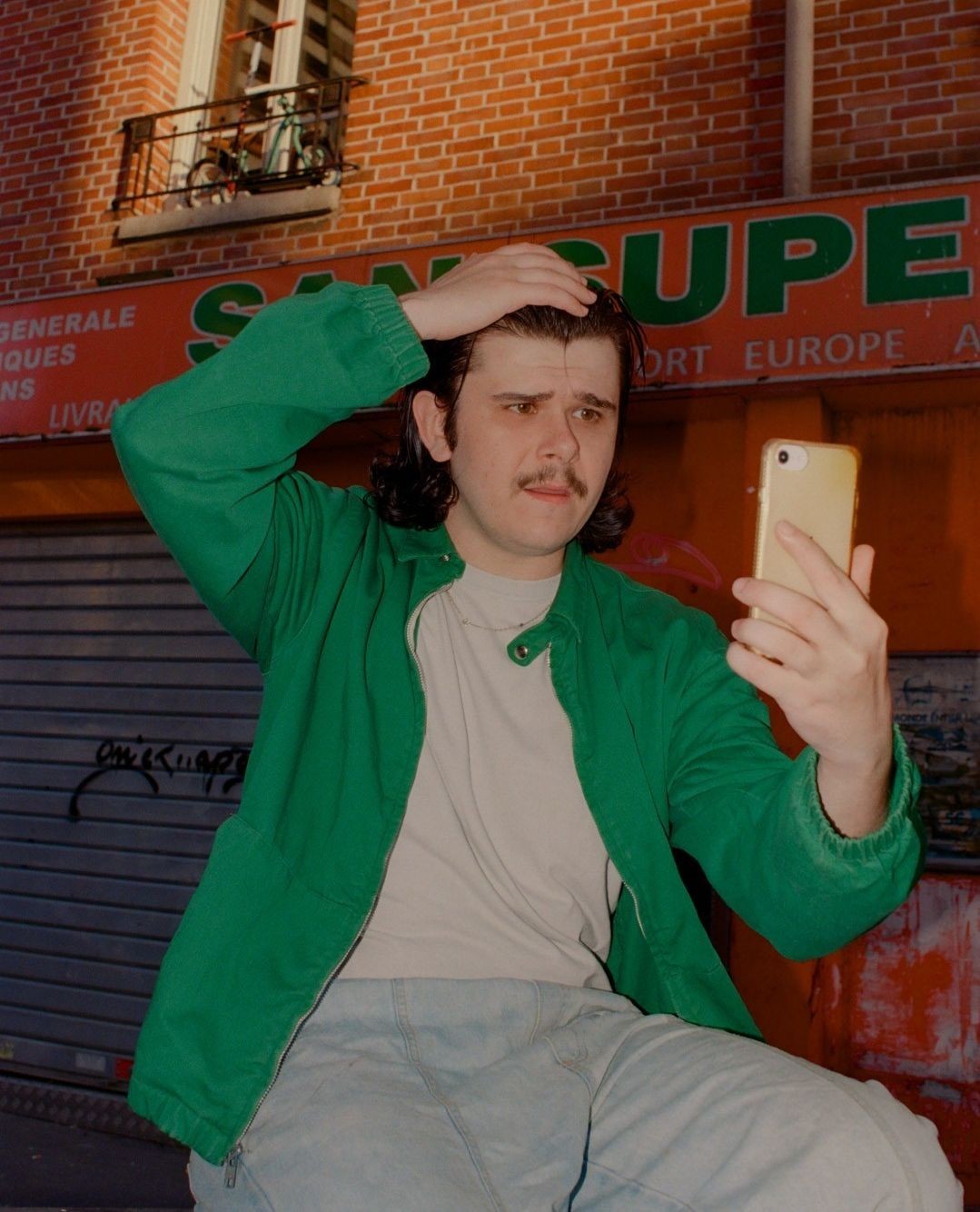 Adult male in a green jacket poses for a phone selfie on a city street with brick buildings and a storefront with European text.