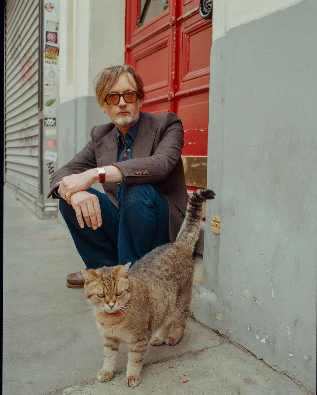 Man with glasses squatting next to a tabby cat on a city street with a red door in the background, posing for a portrait shot.